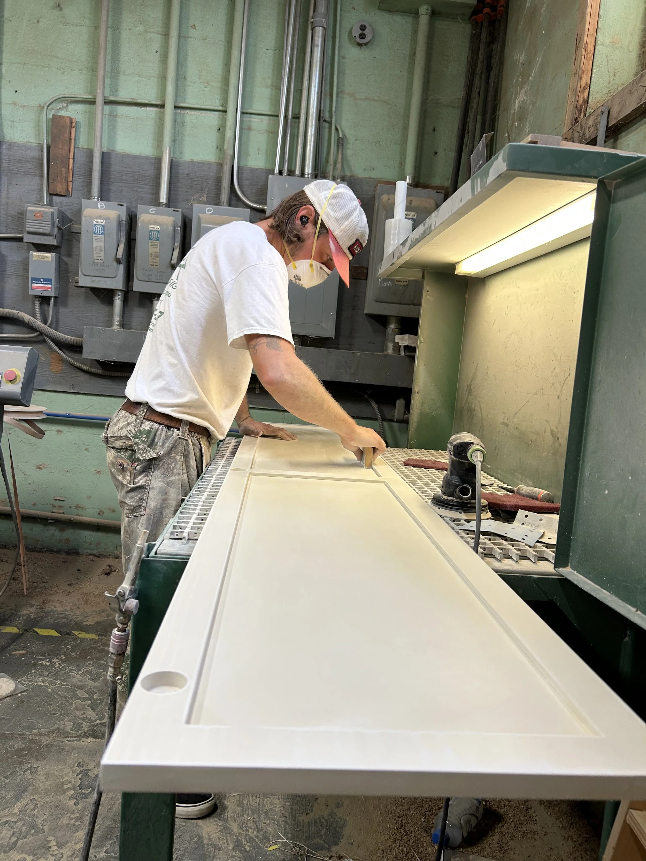 A man working on a woodworking project at a green workbench in a workshop, wearing a white t-shirt, camouflage shorts, a baseball cap, and a mask, using a tool on a large piece of wood.