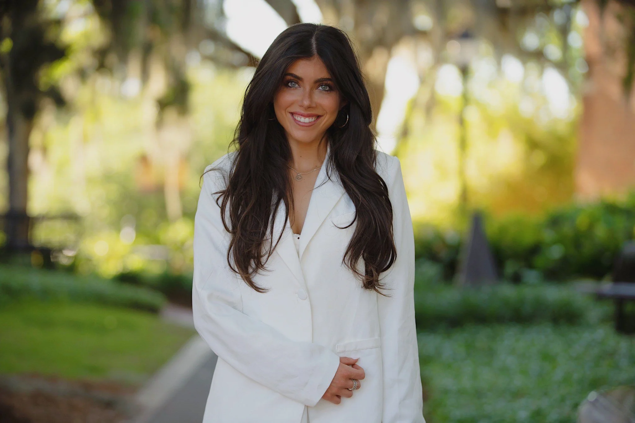Jayden Beir, a therapist with long dark hair wearing a white blazer in a park setting with trees and greenery in the background.