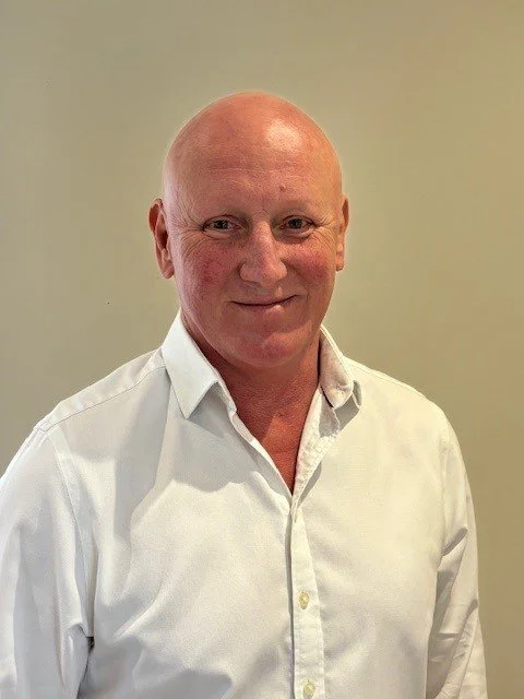 Bald man with a light pink complexion wearing a white dress shirt, smiling, standing against a plain beige wall.