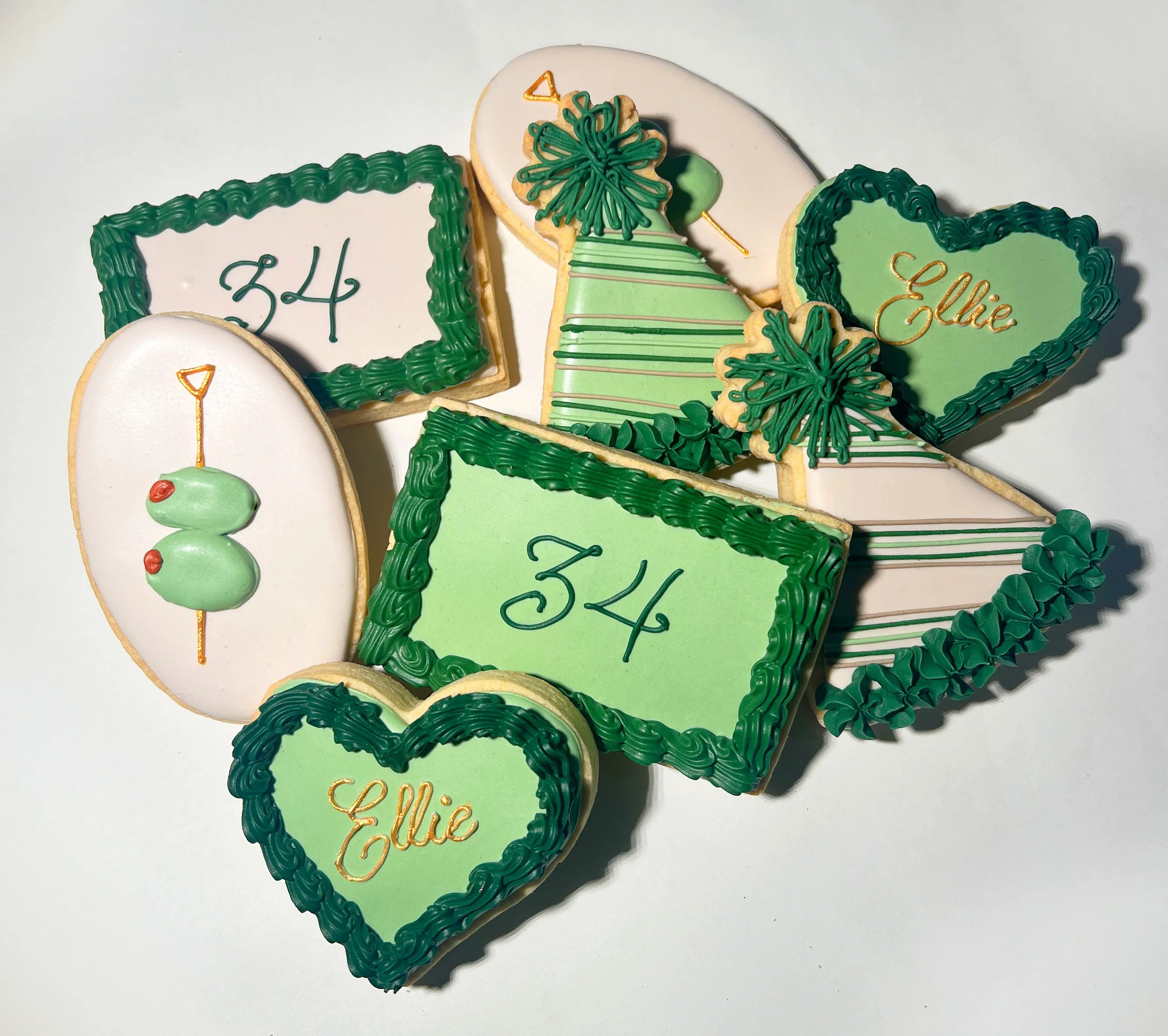Assorted decorated birthday cookies with green and white icing, some with the number 34, others with the name Ellie, and a few with green shamrock and heart shapes.
