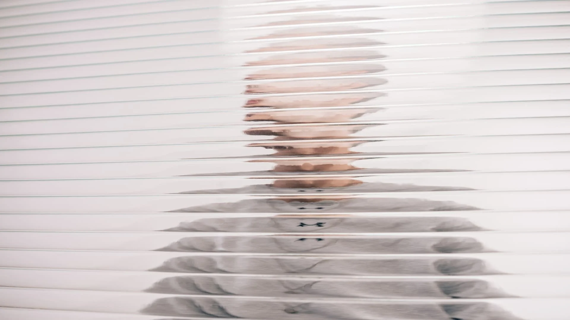 Person standing behind horizontal white blinds, visible through gaps in the blinds.