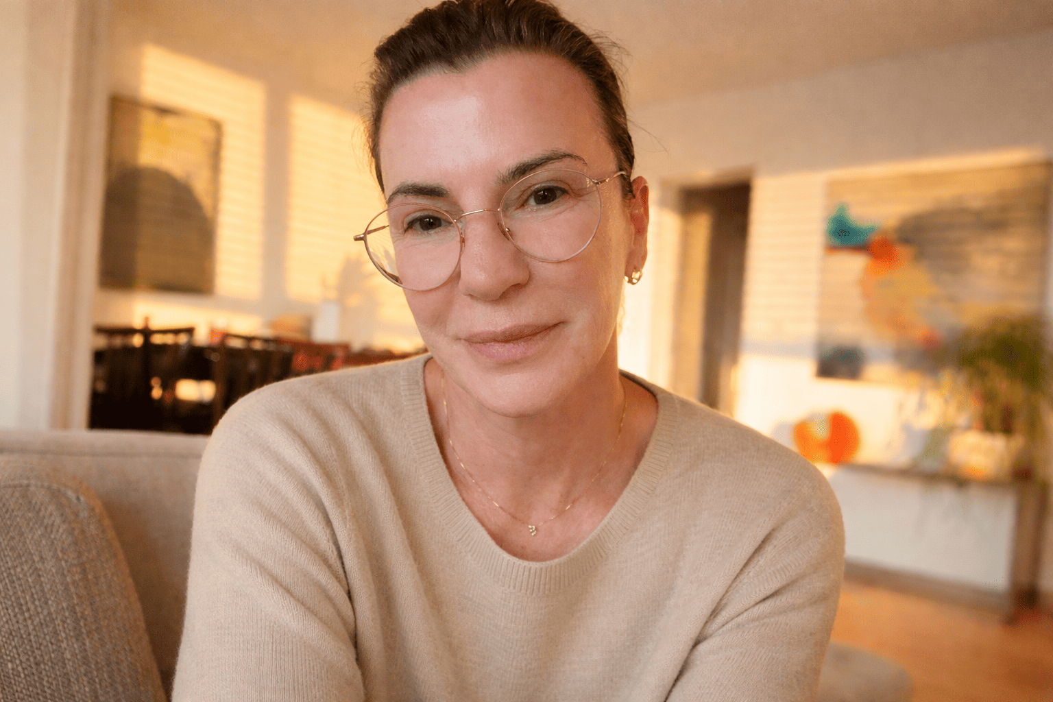 Close-up of a woman with glasses and a beige sweater smiling in a warmly lit living room with artwork and window blinds in the background.