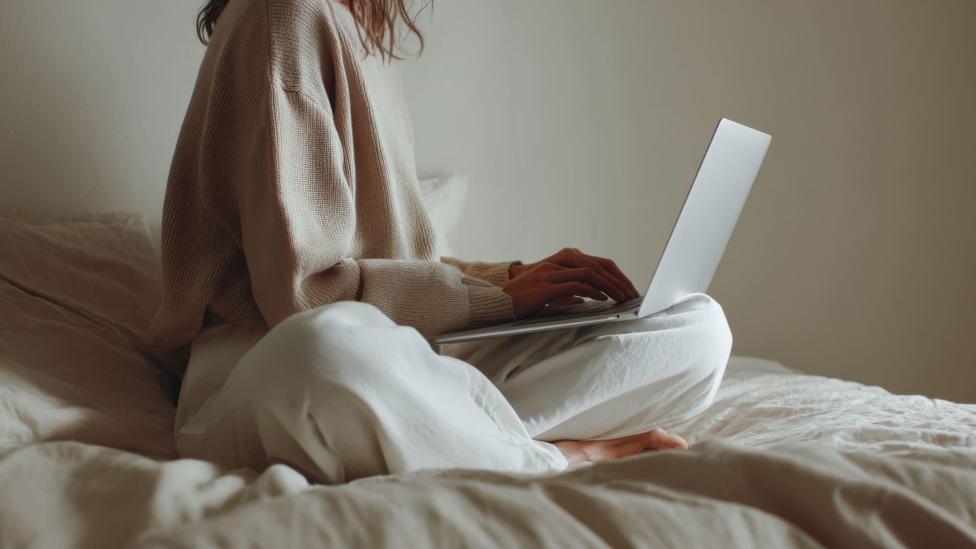 A woman sitting on a bed working on a laptop, wearing beige sweater and white pants, in a softly lit room.