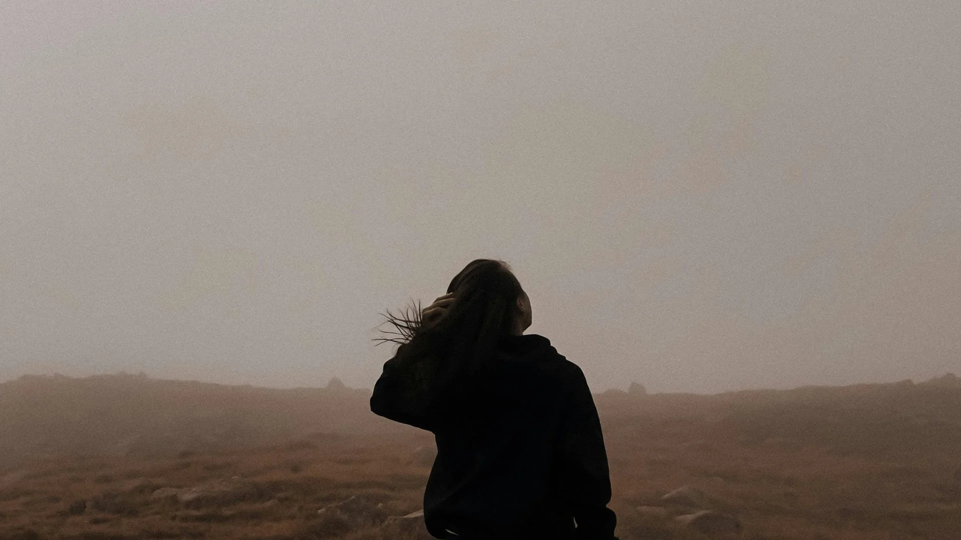 A woman standing outdoors on a cloudy day with her hand behind her head, looking to the side in a barren landscape.