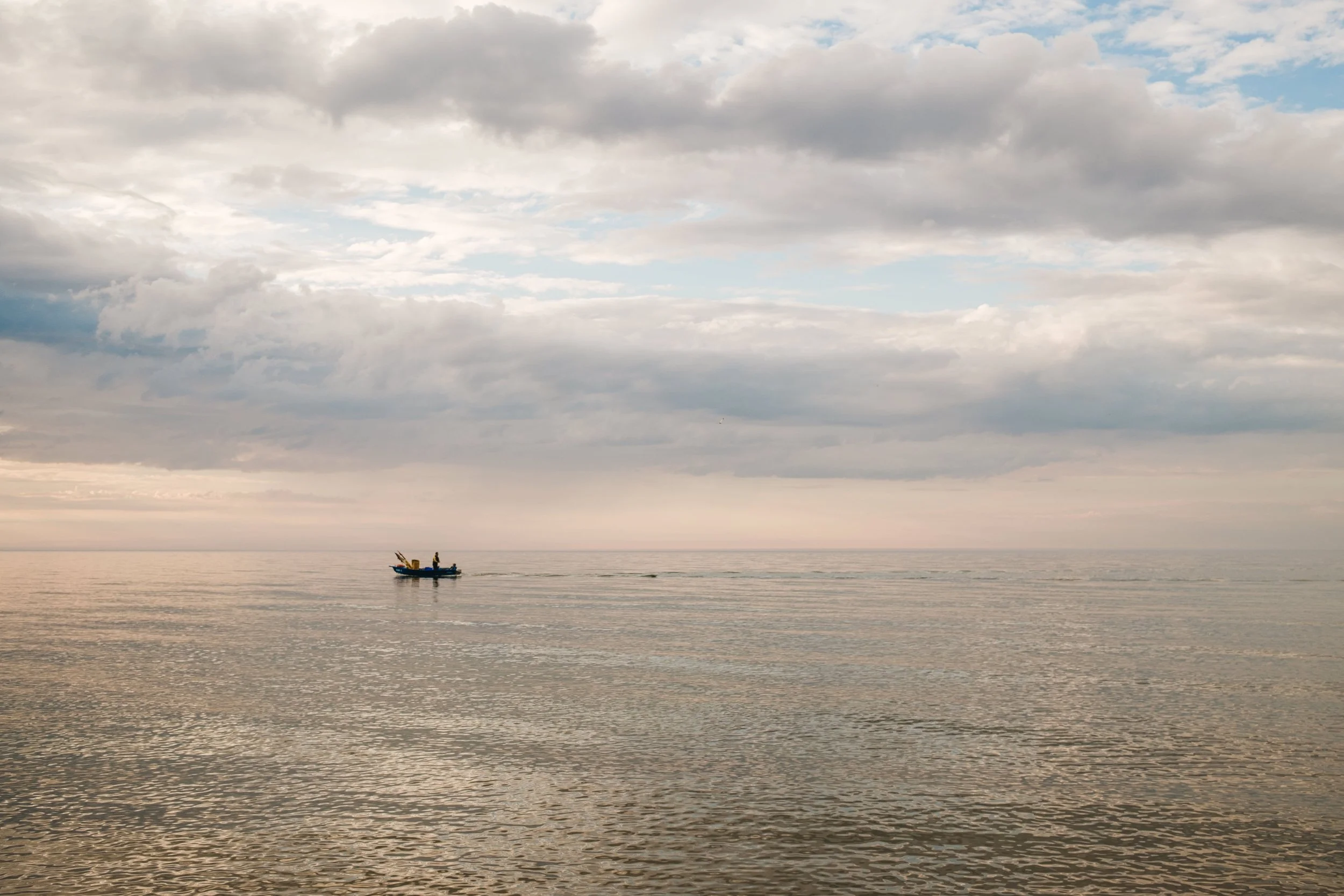 A small boat with two people on calm ocean water under a cloudy sky at sunset.