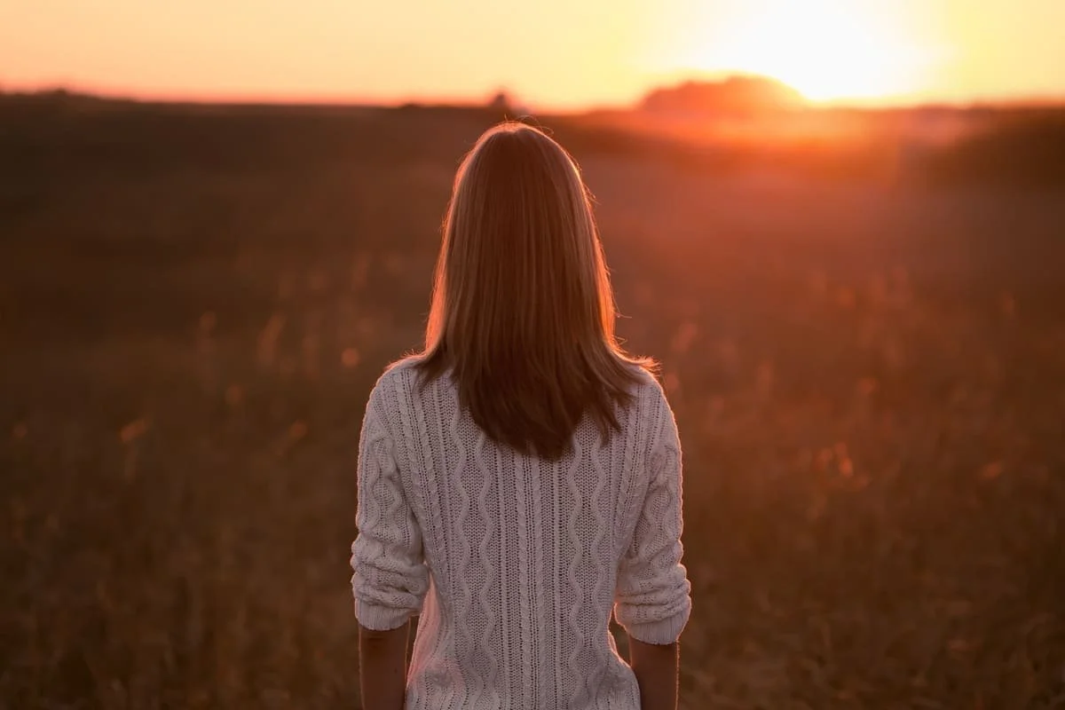 A woman with shoulder-length hair, wearing a knitted sweater, stands outdoors during sunset, facing away from the camera.