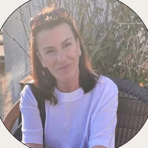 A woman with dark hair and sunglasses on her head sitting outdoors in front of plants, wearing a white shirt.