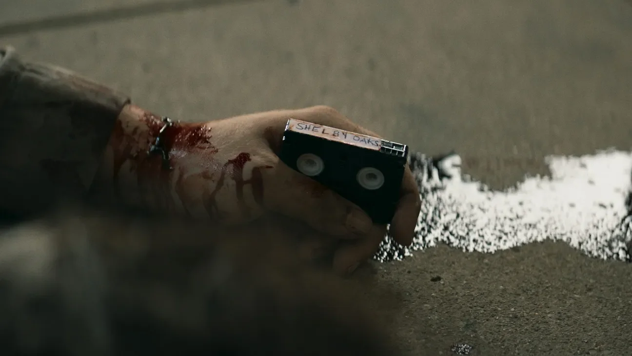 a bloody hand holding a mini VHS tape that has "Shelby Oaks" written on it.