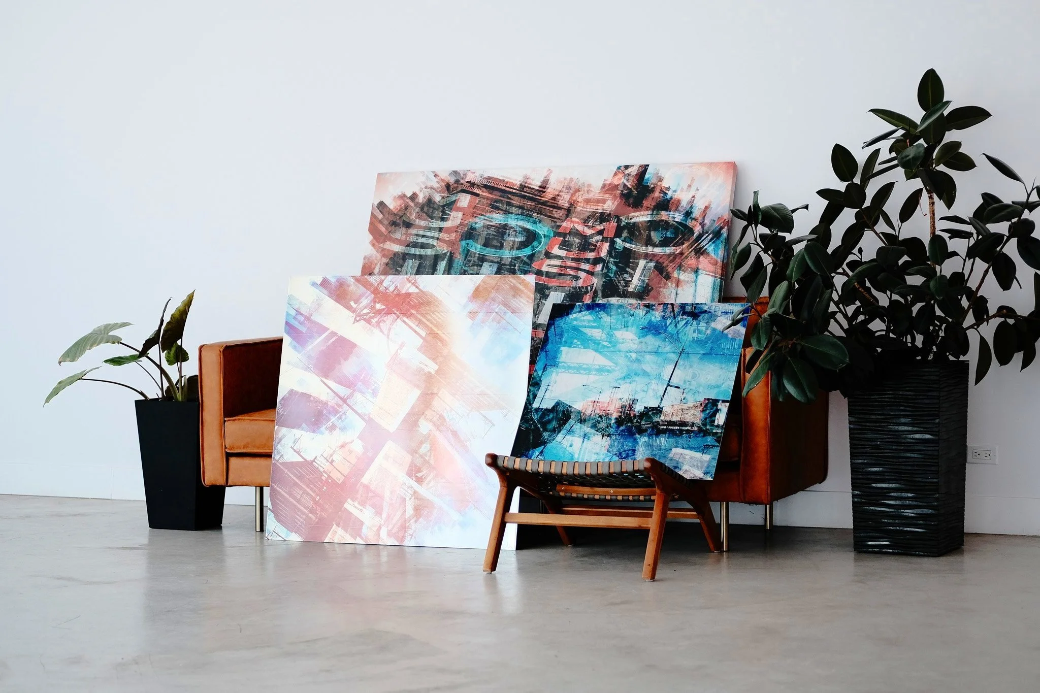 Interior scene with abstract paintings leaning against a white wall, a brown leather chair, a large potted plant, and smaller plants in black pots, with natural light and a power outlet visible.