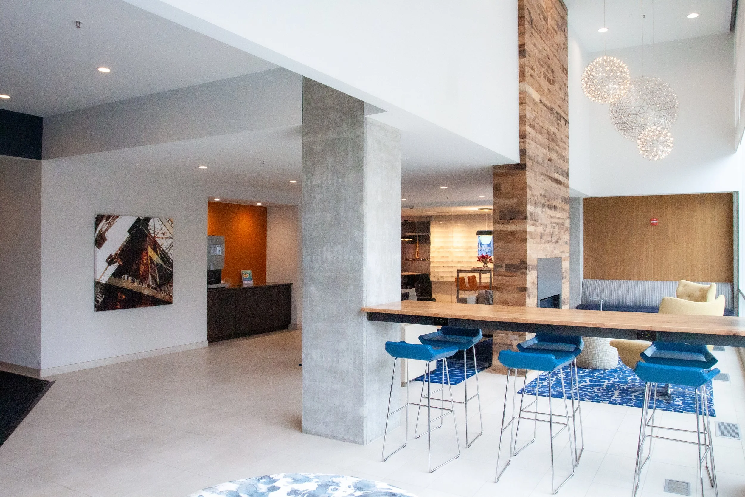 Modern lobby interior with a seating area, a dining table with blue chairs, a large hanging chandelier, a brick wall, and contemporary artwork on a white wall.