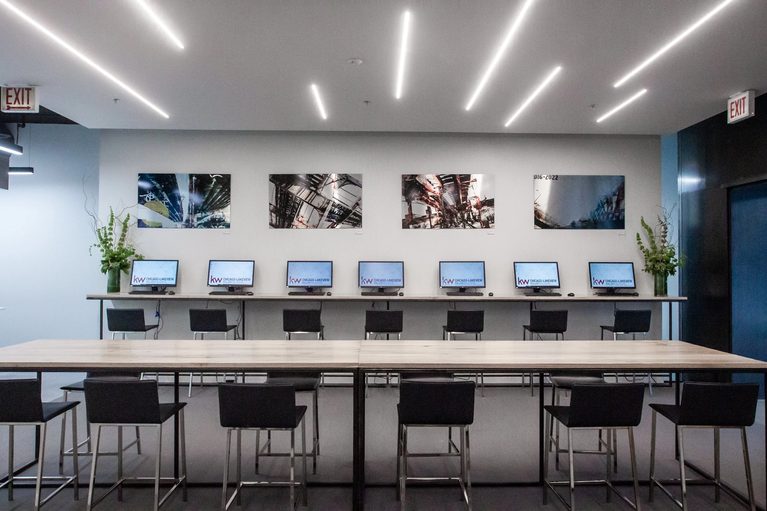 An indoor office space with a row of computers on a long counter, black chairs below, framed artwork on the wall, green plants on each side, and modern ceiling lighting.