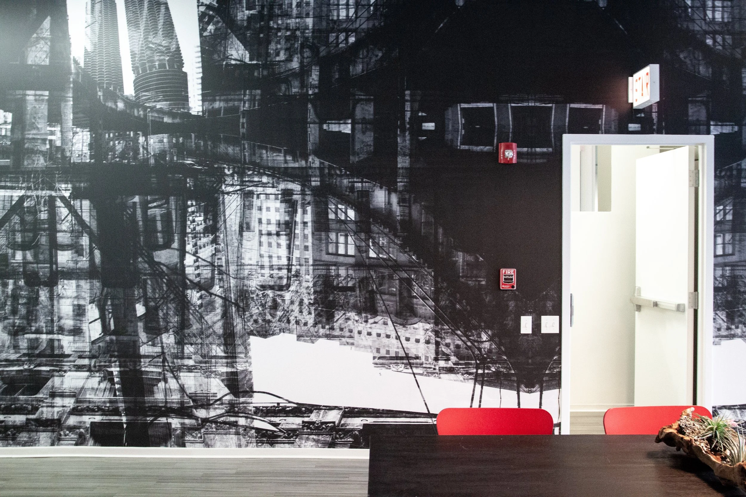 Interior of a room with a black and white urban cityscape mural on the wall, a white door slightly open leading to another room, a fire alarm and extinguisher on the wall, two red chairs, and a table with a decorative object.
