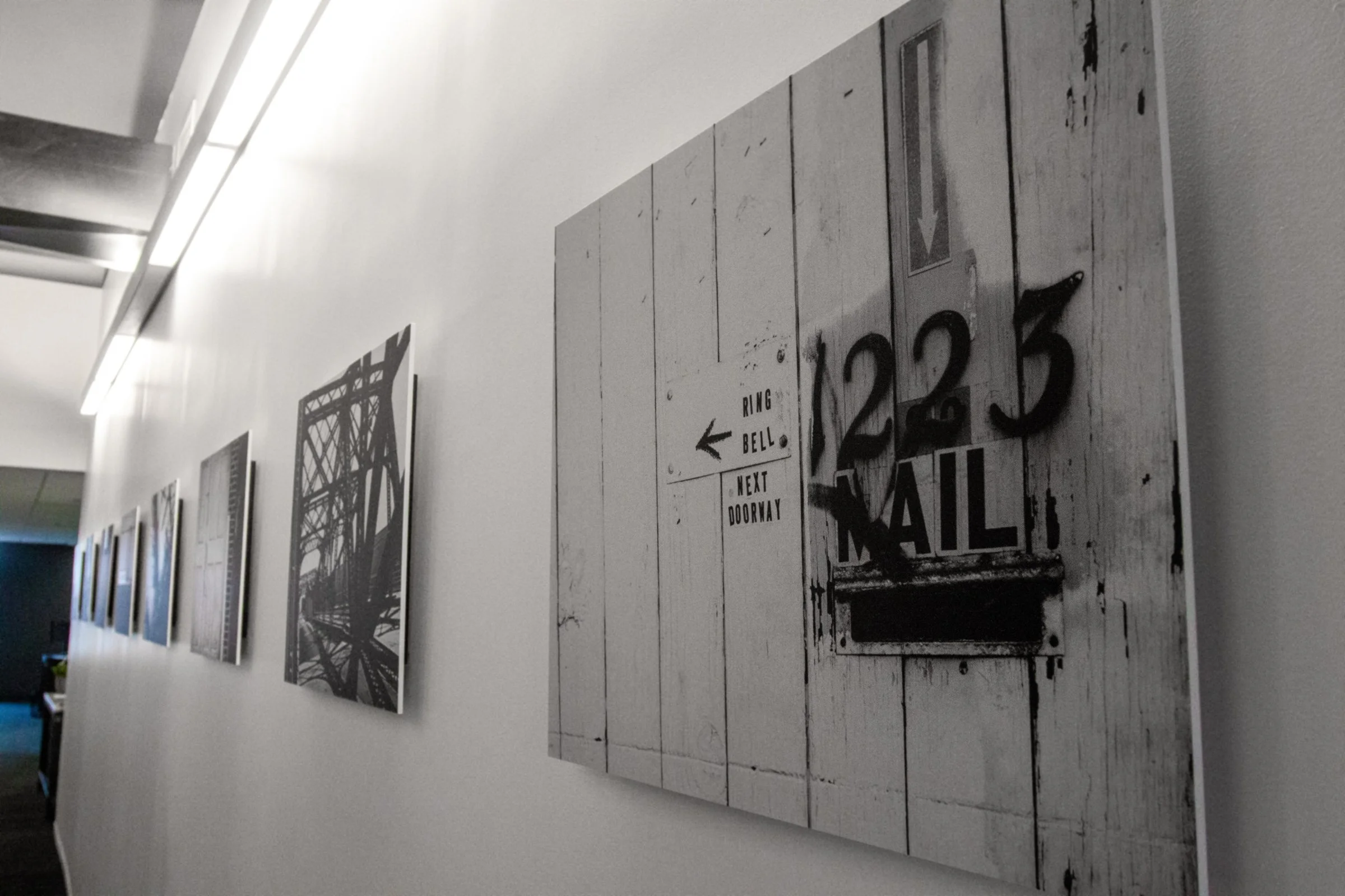 Black and white artwork featuring a bridge structure and signage with directions, mounted on a white wall in an art gallery.