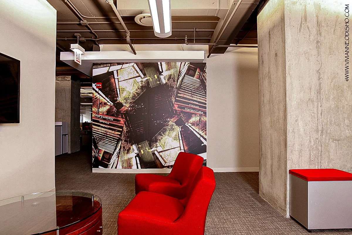 Modern office lobby with a red chair, a small table, and a wall with a large abstract photo mural of a cityscape and train tracks.