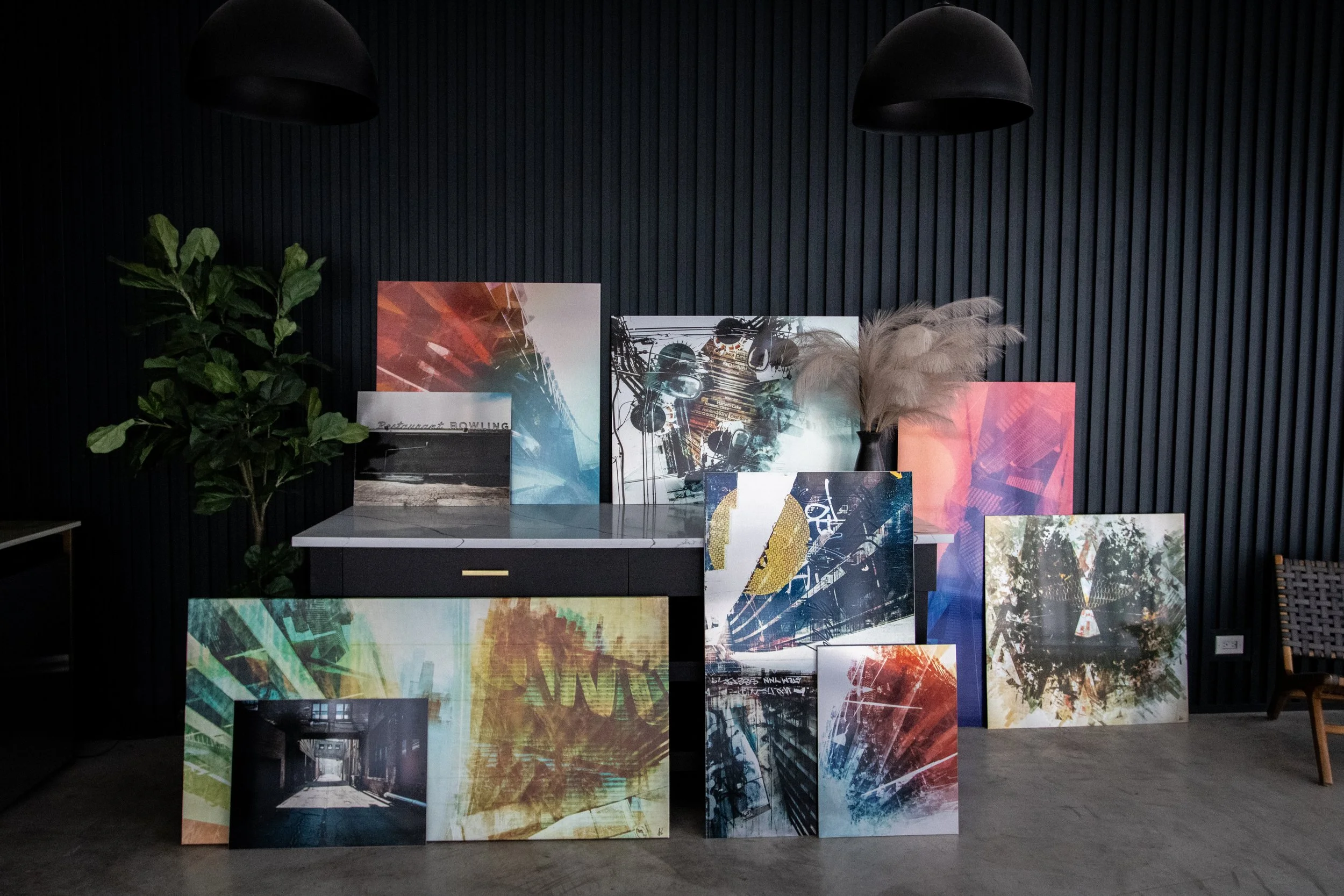 An indoor display of various abstract colorful art prints arranged against a black paneled wall, with a green potted plant on the left and a dark chair on the right.