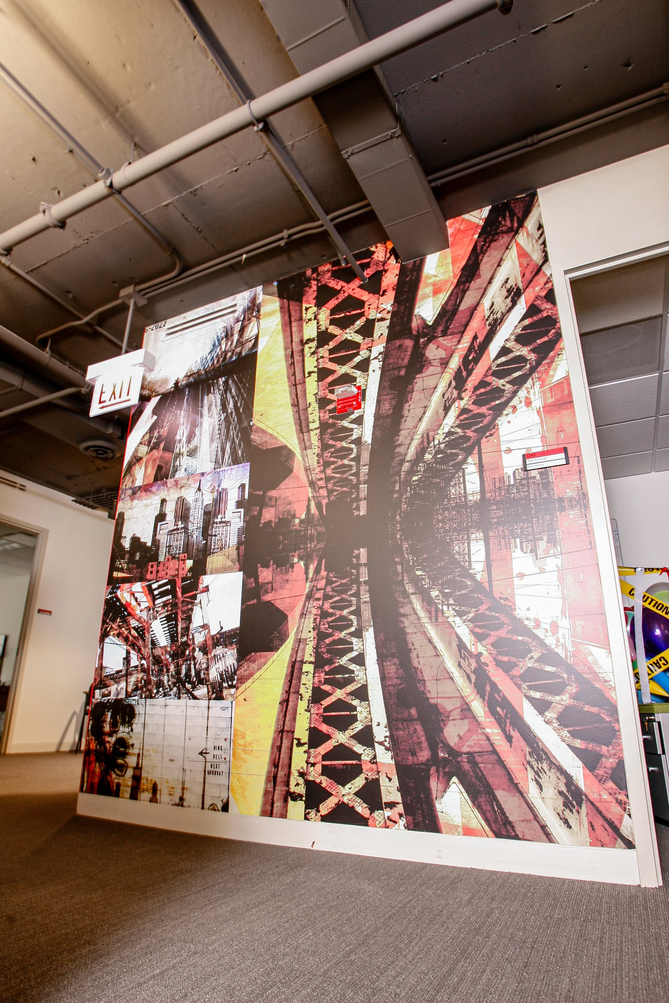 An indoor art installation featuring a large mural of a bridge, cityscape, and urban elements with a red and black color scheme, hanging on a wall with exposed ceiling pipes.