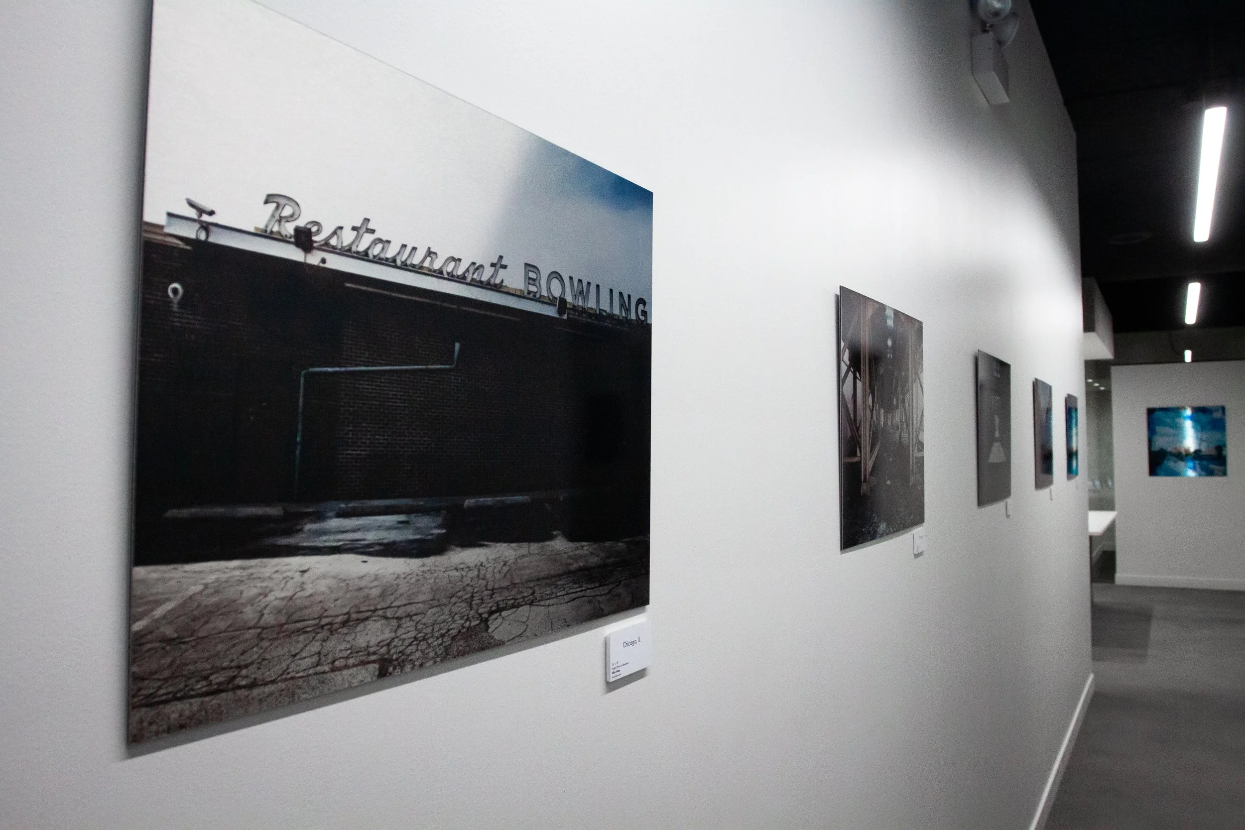 Photographs displayed on a gallery wall, including a photo of a building with a neon sign that says 'Restaurant Bowling' and a dark, cracked pavement near the building, with other framed images further down the hallway.