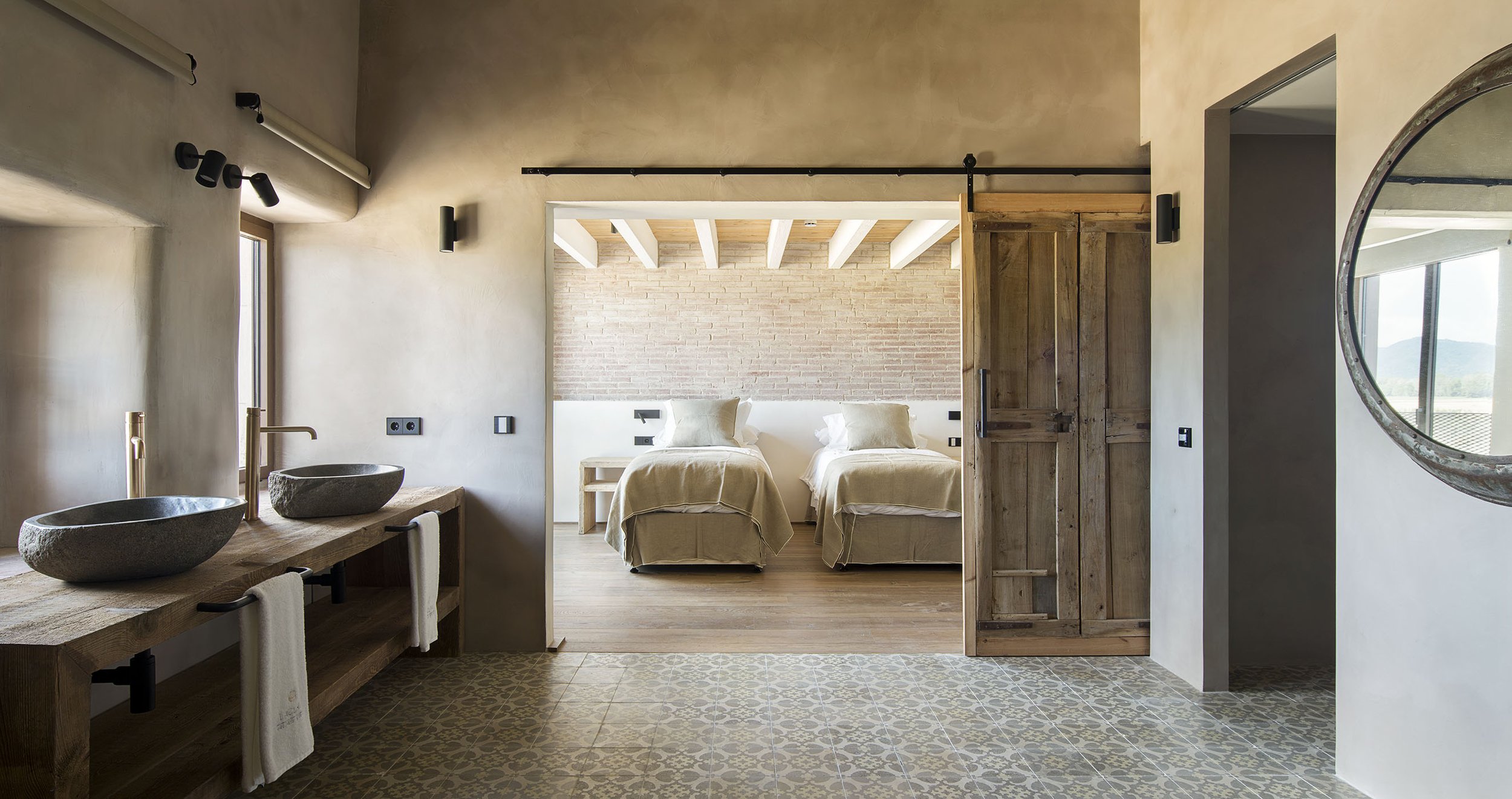 A bedroom with patterned wallpaper, a mirror, flat-screen TV, and mounted rifles. The bed has green pillows, a beige pillow, and a green blanket. There are two bedside lamps, a wooden desk with a chair, and a radiator.