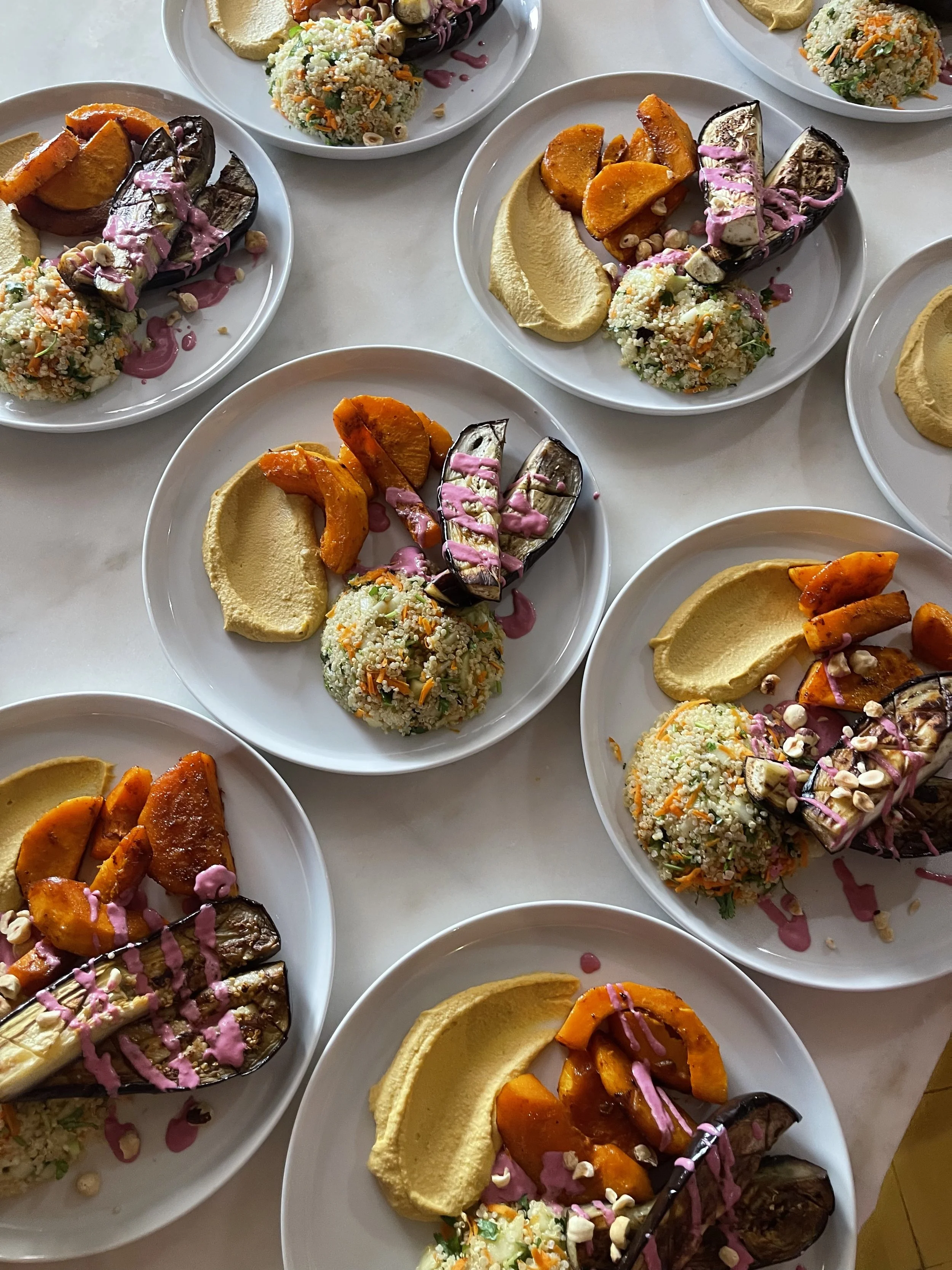 Plates of roasted eggplant, roasted sweet potatoes, hummus, and a grain salad with vegetables and nuts.