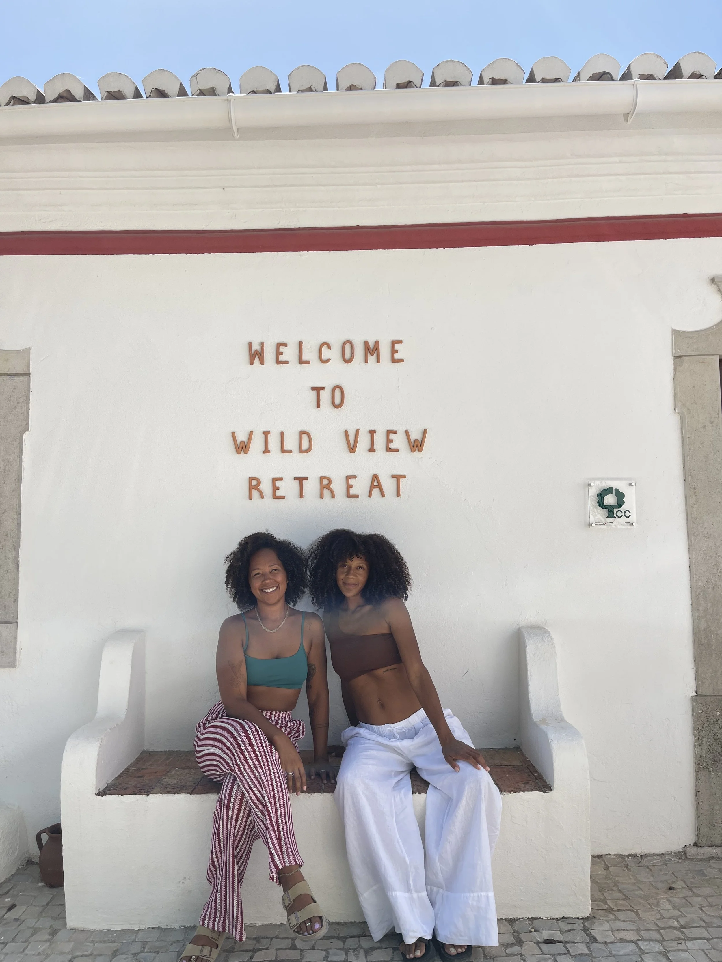 Two black women sitting on a white bench in front of a white wall with the sign 'Welcome to Wild View Retreat.' They are smiling, with curly hair, wearing tank tops and wide-legged pants, and appear happy, on a yoga retreat in Portugal.
