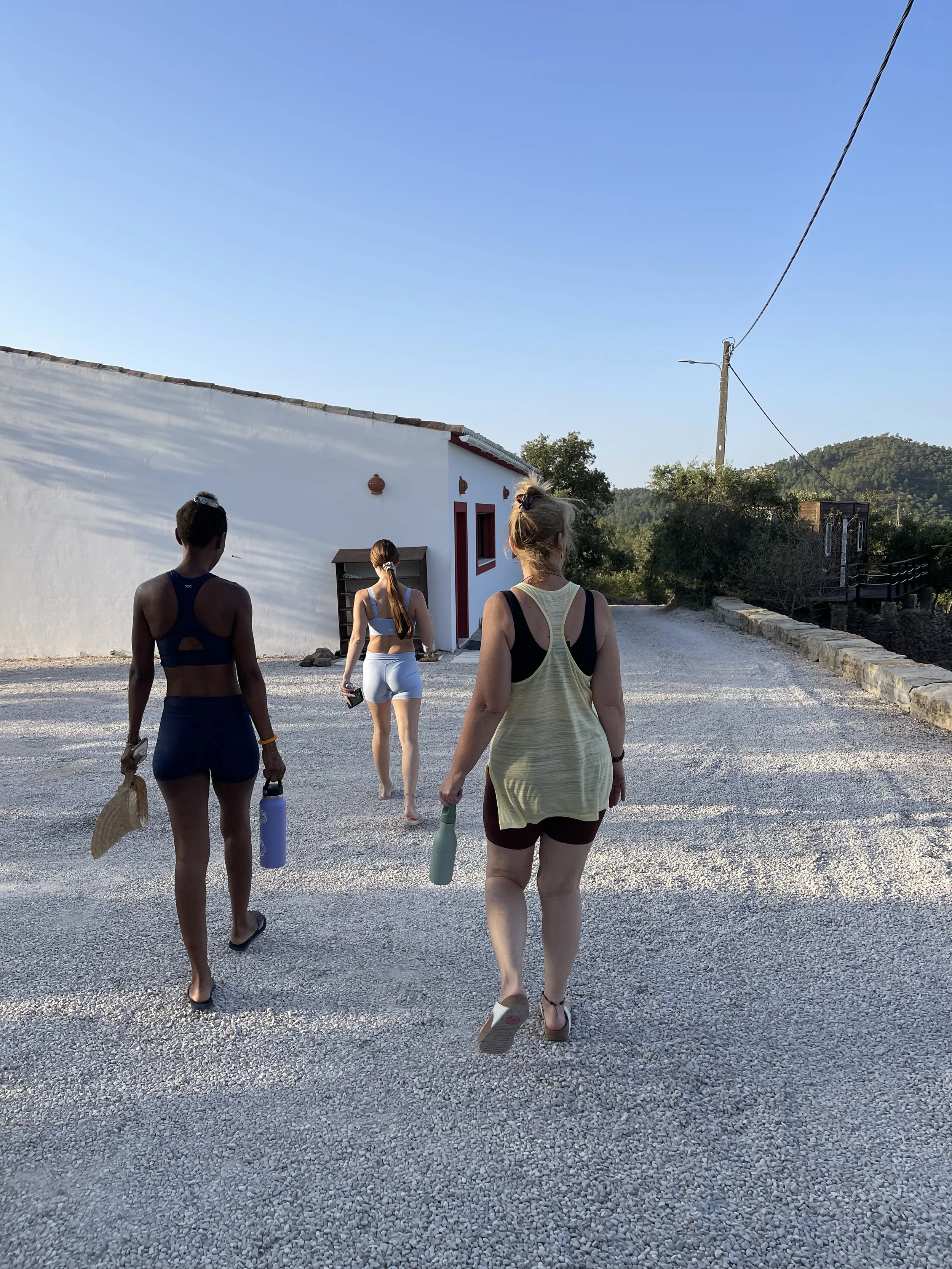 Three women dressed in athletic wear walking down a gravel path toward a white building with red accents, with hills and trees in the background on a sunny day, on a yoga retreat in Portugal.