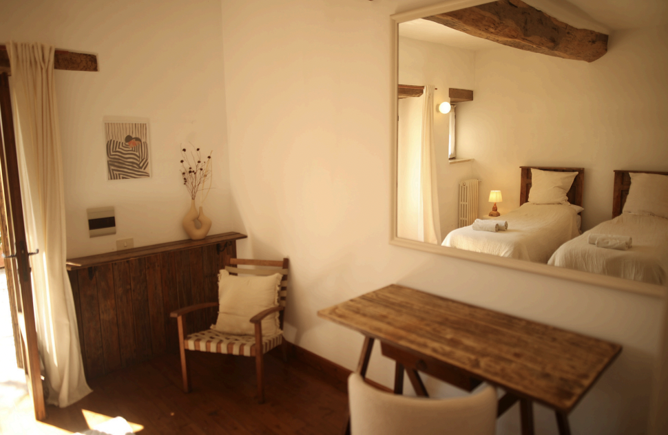 A cozy bedroom with a large bed, beige headboard, multiple pillows, bedside lamp, wooden side table, and sheer curtains allowing natural light.