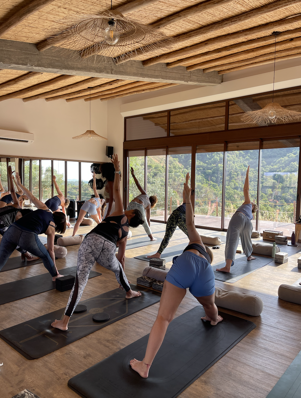 A group of people participating in a yoga class in a bright room with wooden floors, large windows, and scenic outdoor views. They are performing side stretches on mats, with some using yoga props like blocks and bolsters, on a yoga retreat in Portu