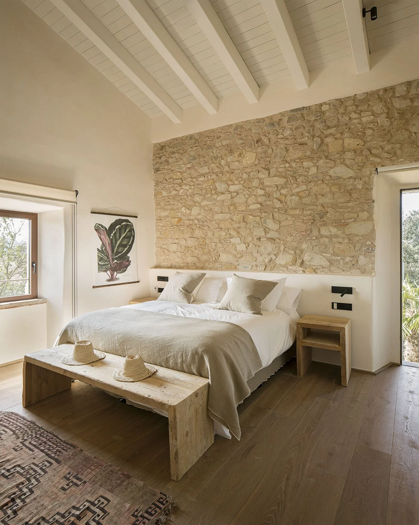 A cozy bedroom with a large bed featuring a rustic wooden headboard, white bedding, and black and white pillows. There are nightstands with lamps on each side of the bed, a sliding glass door with curtains, and a standing blue lamp in the foreground. A wooden door and a partial view of a sitting area are also visible.