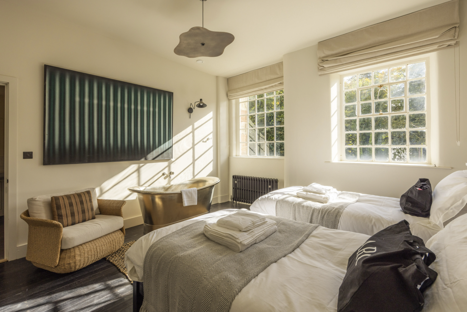 A bedroom with patterned wallpaper, a mirror, flat-screen TV, and mounted rifles. The bed has green pillows, a beige pillow, and a green blanket. There are two bedside lamps, a wooden desk with a chair, and a radiator.