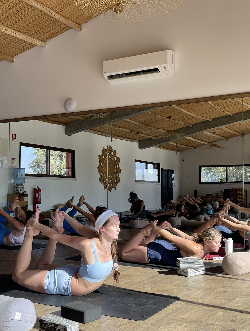 A group of people practicing yoga in a bright, spacious studio with wooden ceiling beams, windows, some meditation cushions, and yoga mats, with a woman in the foreground wearing a light blue sports bra and headband, on a yoga retreat in Portugal.