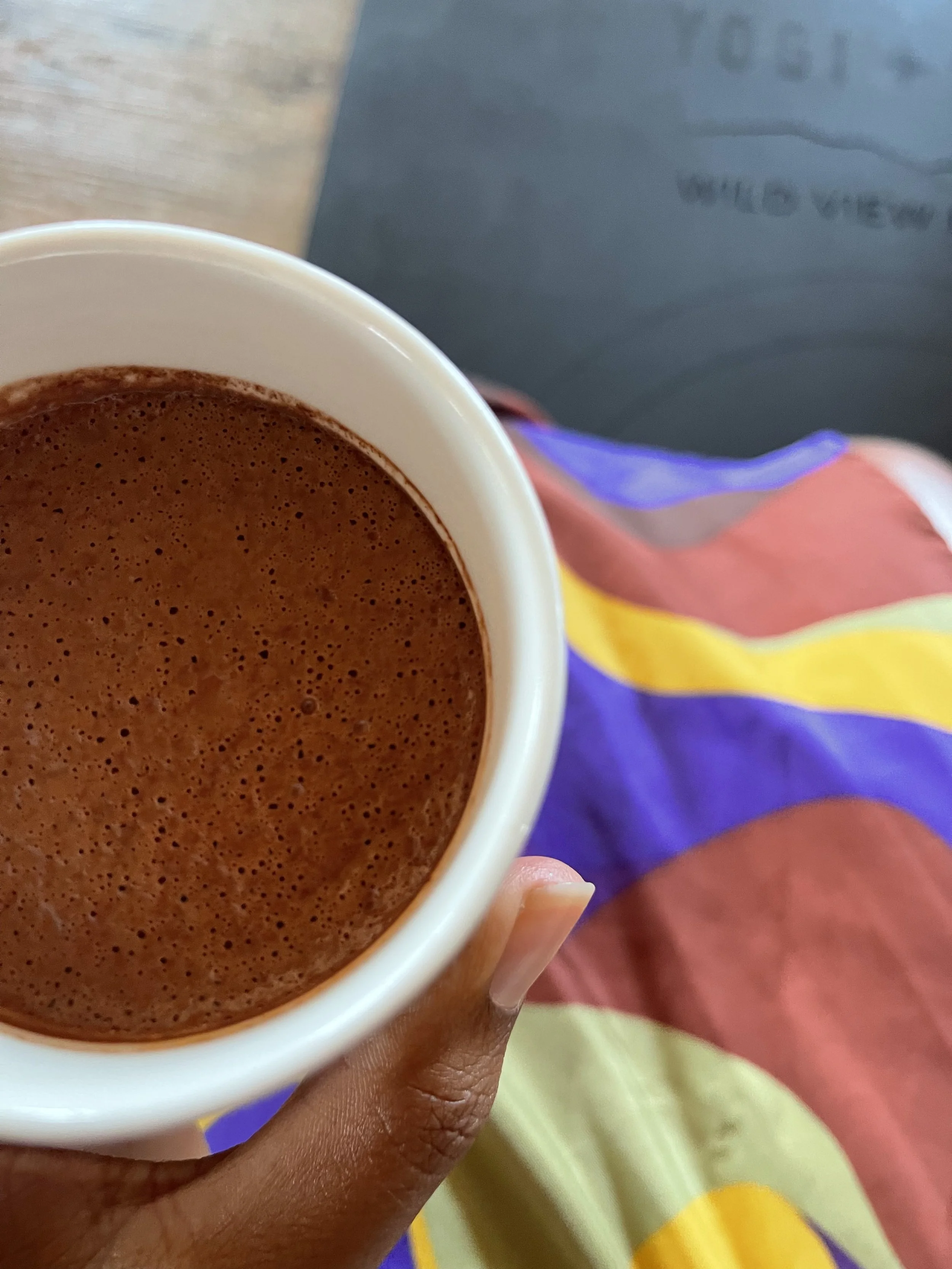 A close-up of a hand holding a white mug filled with hot chocolate or coffee. Background includes colorful fabric with purple, yellow, red, and green colors, and a black surface with some blurry text.