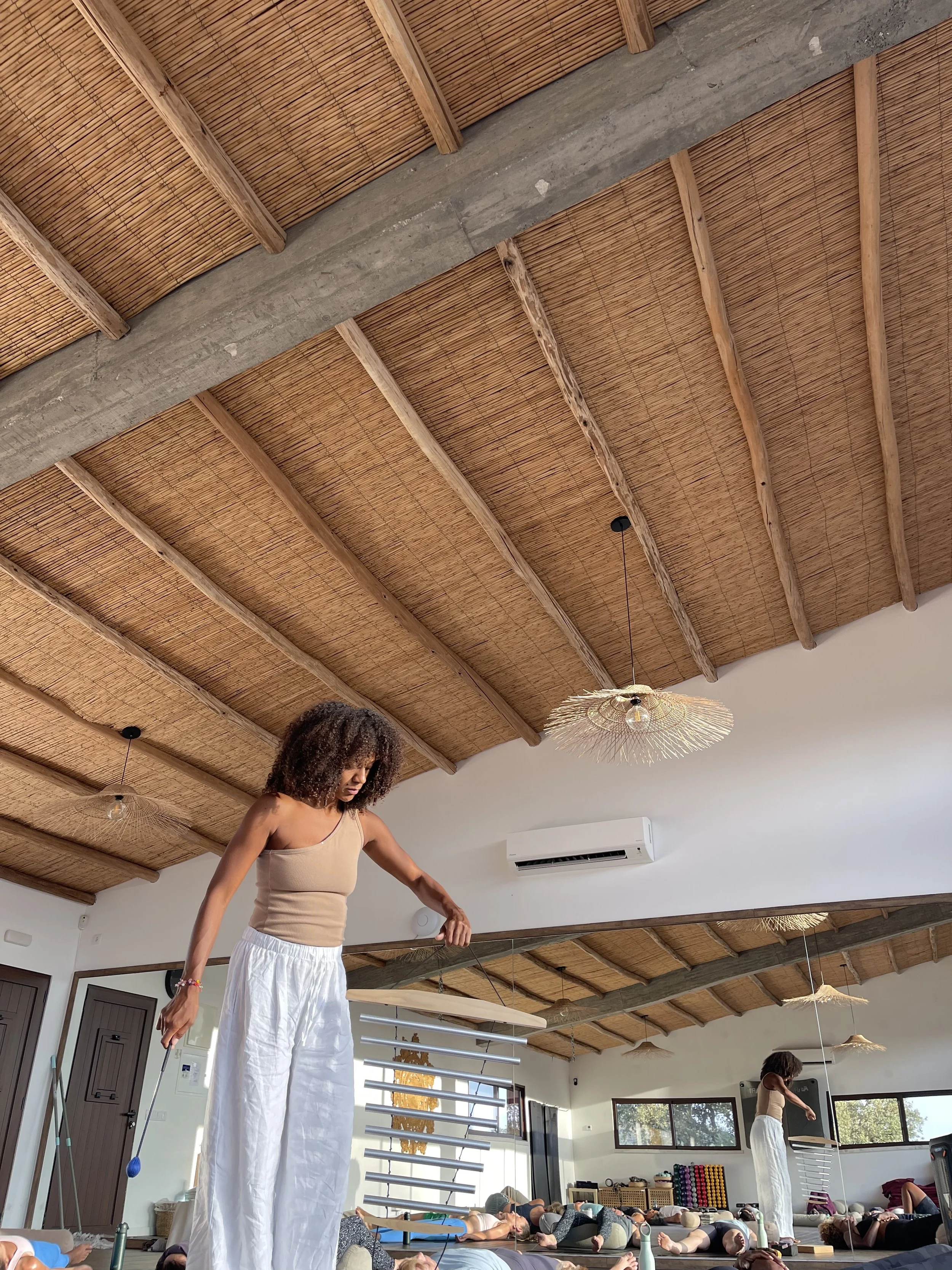 A black woman leading a yoga class in a spacious studio with a thatched wood ceiling, practicing yoga with students lying on mats, on a yoga retreat in Portugal.