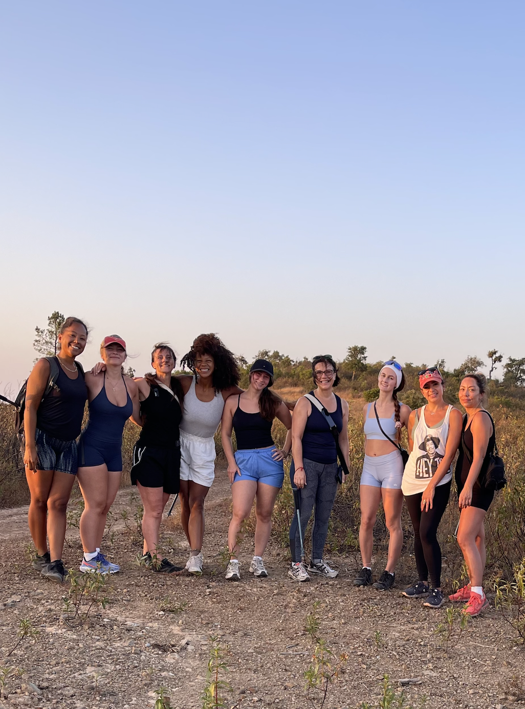 A group of nine women standing outdoors on a dirt trail, smiling, with dry grass and trees in the background during sunset.