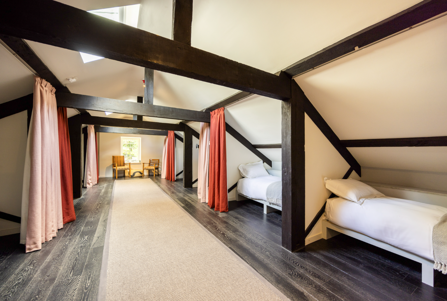 A cozy bedroom with a large bed featuring a rustic wooden headboard, white bedding, and black and white pillows. There are nightstands with lamps on each side of the bed, a sliding glass door with curtains, and a standing blue lamp in the foreground. A wooden door and a partial view of a sitting area are also visible.