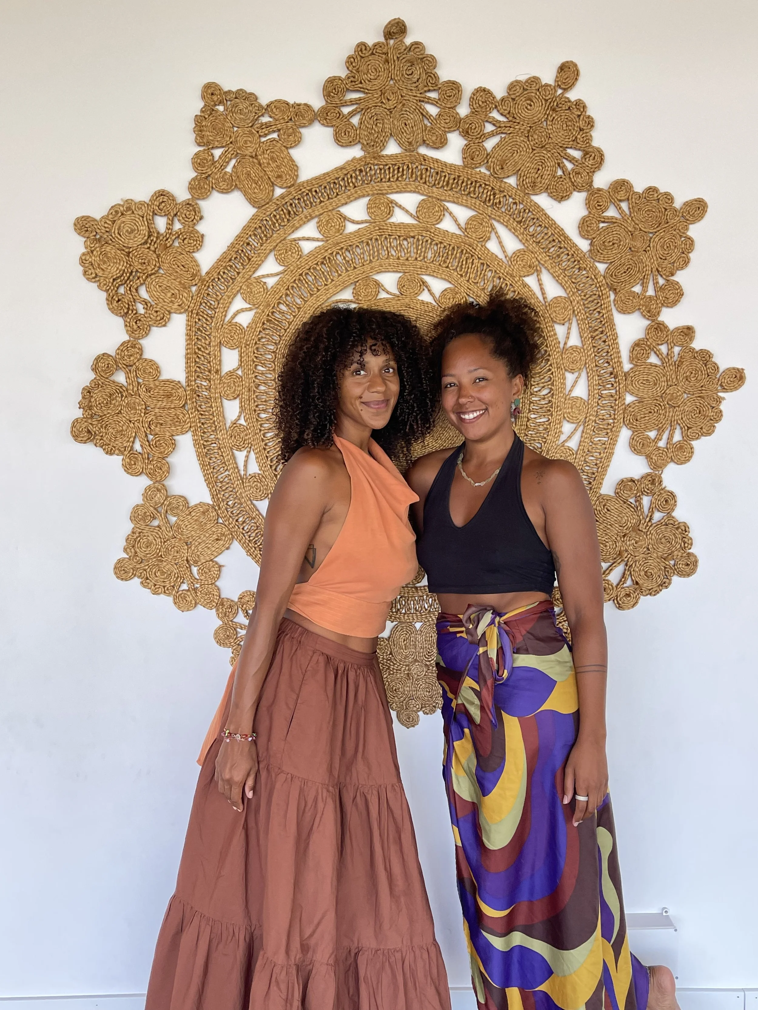 Two black, mixed raced women smiling and standing together in front of an intricate golden wall hanging, on a yoga retreat in Portugal