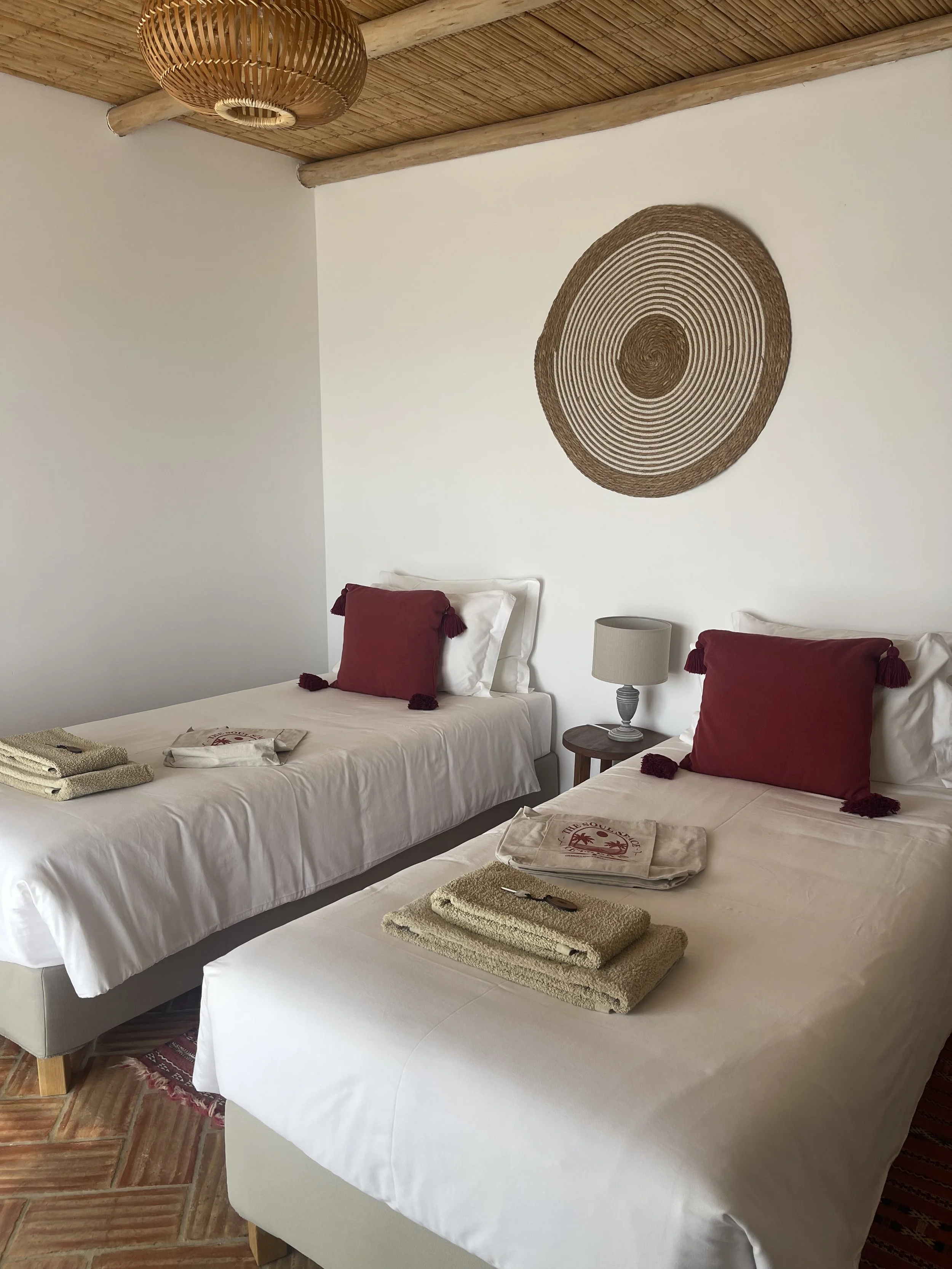 Two neatly made beds with white linens and large red pillows with tassels, a small round bedside table with a lamp, towels on the beds, and a decorative woven wall art piece on a white wall in a room with a wooden ceiling.