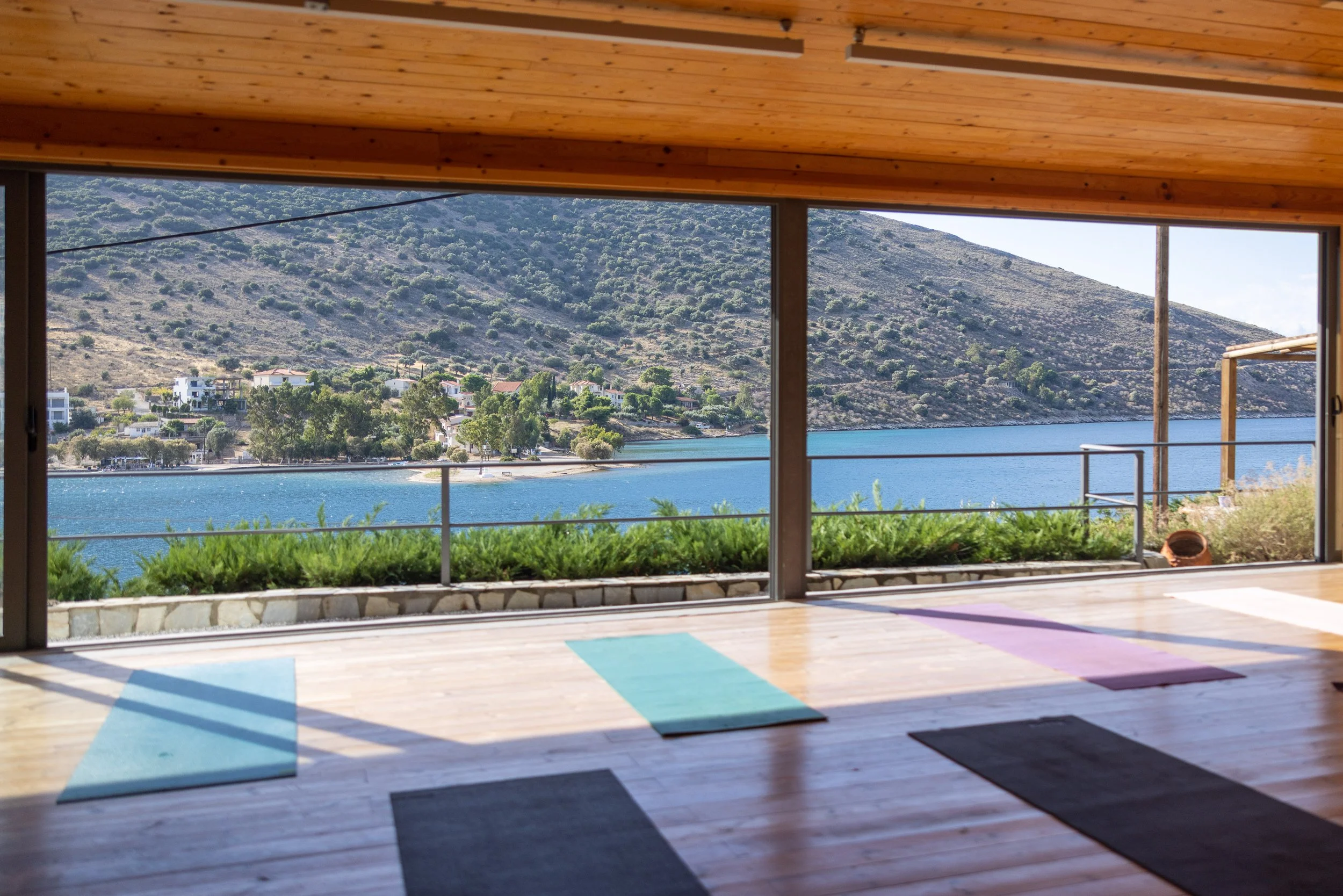 Indoor yoga studio with green mats and blue blocks, large windows showing a lake and mountains outside in Greece.