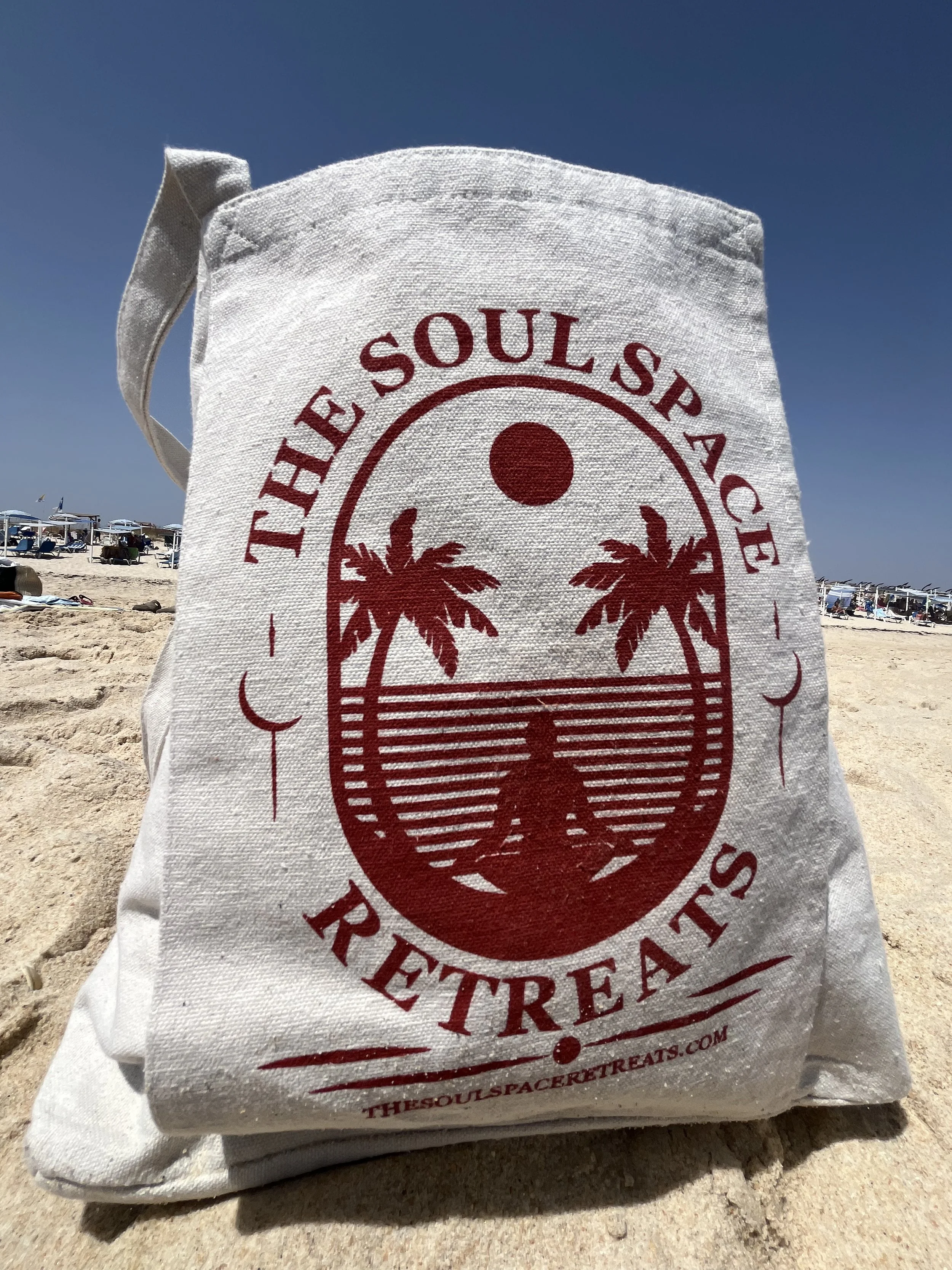 A white tote bag with red print, featuring a design of two palm trees, a person sitting on the ground, a sun, and lines representing water or sunset, with the text 'The Soul Space Retreats' and a website address, on a yoga retreat in Portugal.