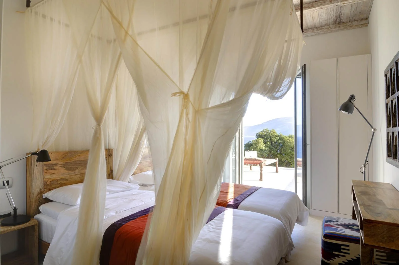 A cozy bedroom with a large bed, beige headboard, multiple pillows, bedside lamp, wooden side table, and sheer curtains allowing natural light.