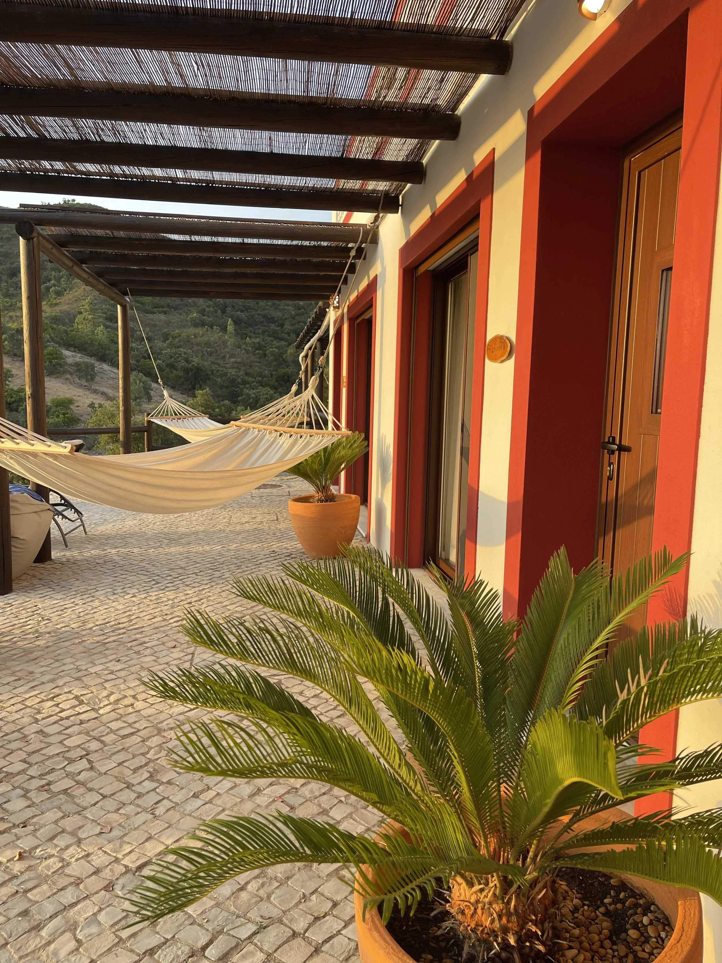 A balcony with a hammock, potted plants, and a view of green hills in the background, during sunset, on a yoga retreat in Portugal.