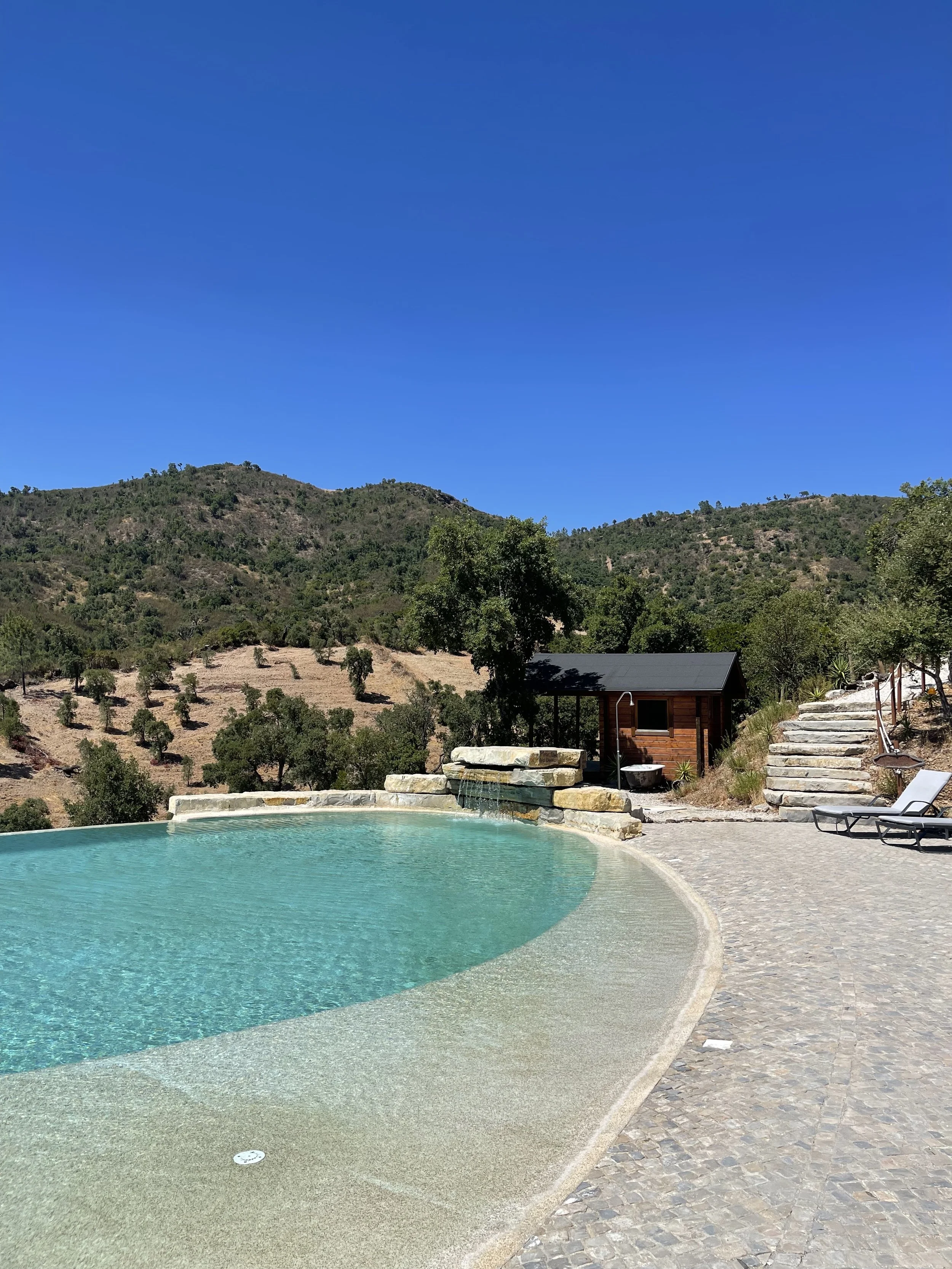A scenic outdoor pool with a mountain view, surrounded by a stone patio and lounge chairs. There is a small wooden shed and steps leading up a hillside, with trees and hills in the background, under a clear blue sky, on a yoga retreat in Portugal.