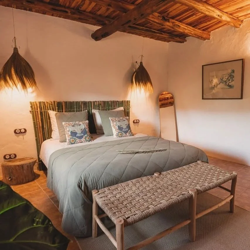 A cozy hotel room with a large bed, beige pillows, wood headboard, hanging woven lamp, curtains covering a balcony door, potted plant, and a white dresser with a kettle and cups.