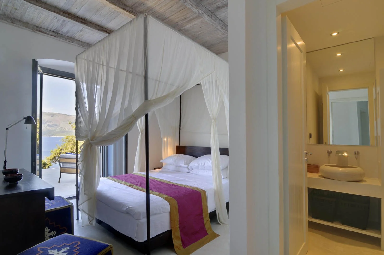 A cozy hotel room with a large bed, beige pillows, wood headboard, hanging woven lamp, curtains covering a balcony door, potted plant, and a white dresser with a kettle and cups.