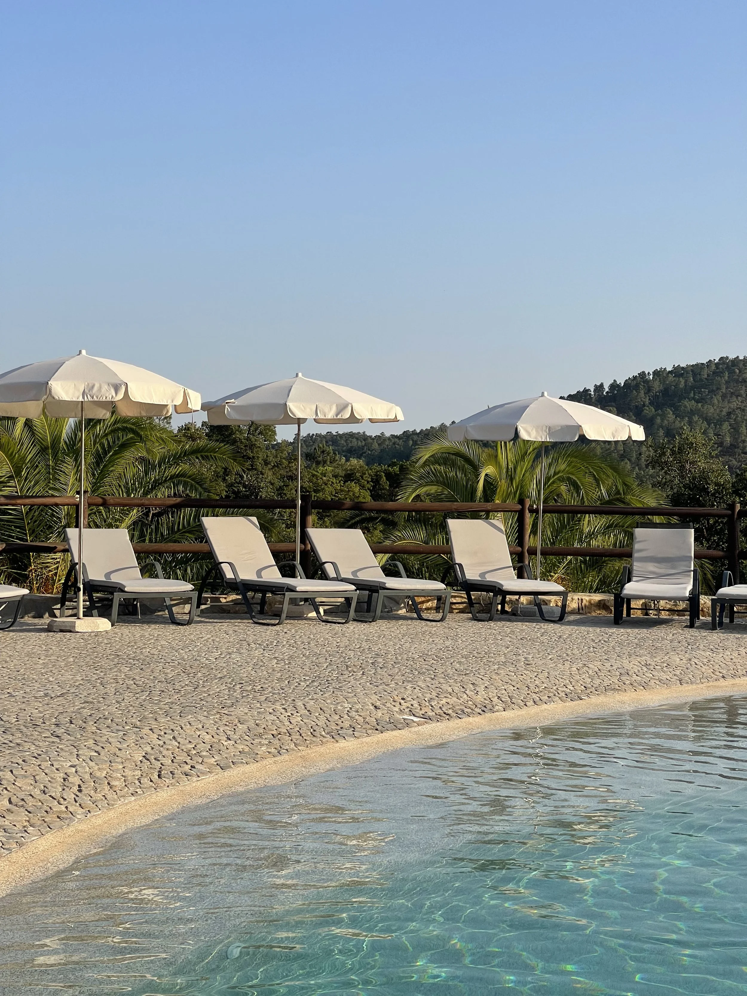 Poolside view with lounge chairs and white umbrellas, lush greenery and hills in the background, clear blue sky, on a yoga retreat in Portugal.