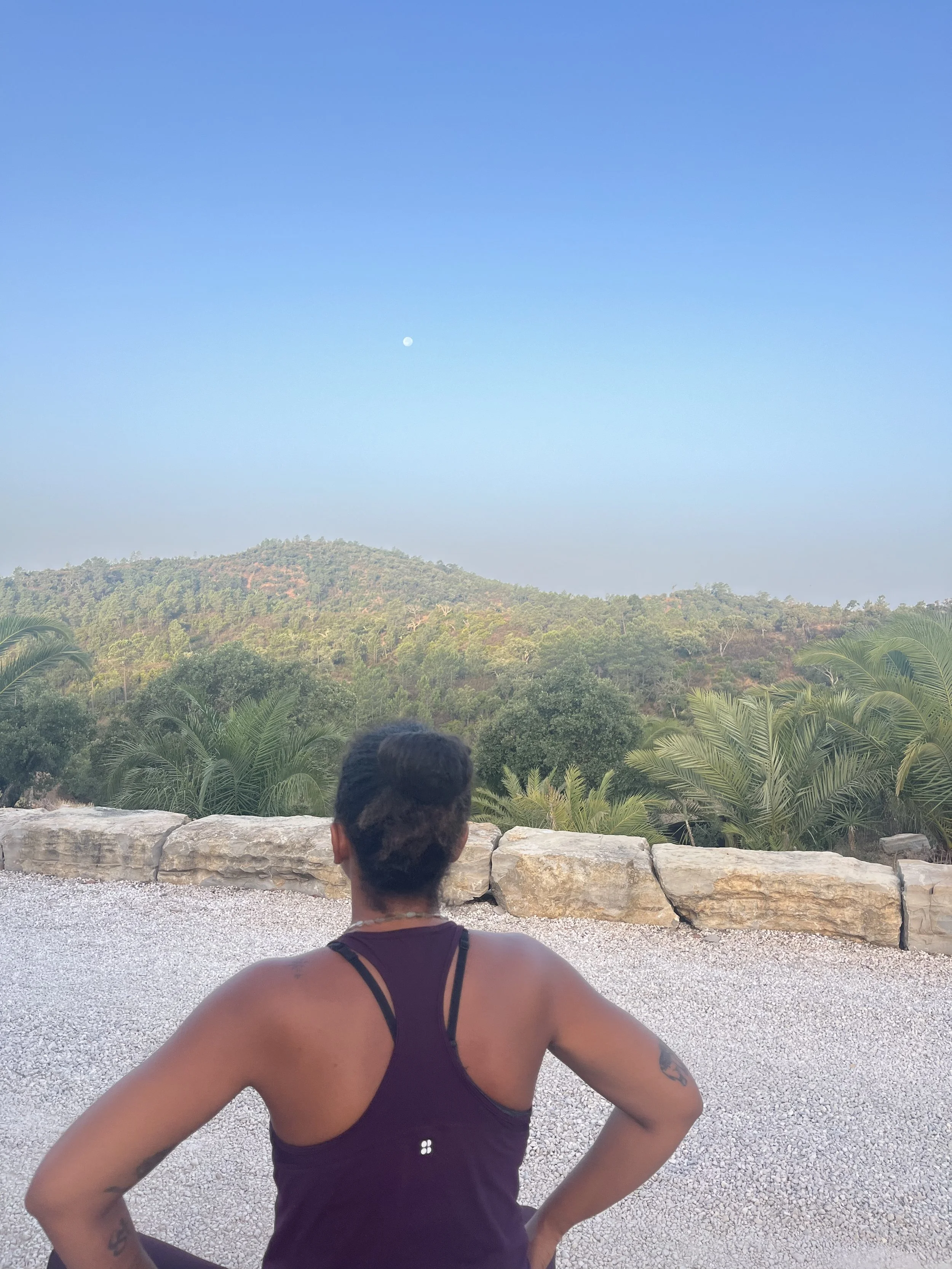 Person with short hair sitting on a stone ledge, overlooking a lush green landscape with trees and hills, and a clear sky with the moon visible, on a yoga retreat in Portugal.