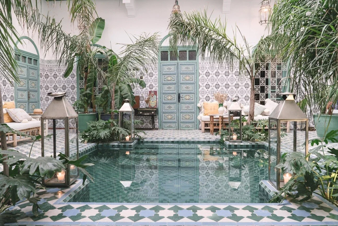 Indoor courtyard with a swimming pool, lush green plants, tiled walls and floor, teal doors, sofas with cushions, and lanterns with candles.