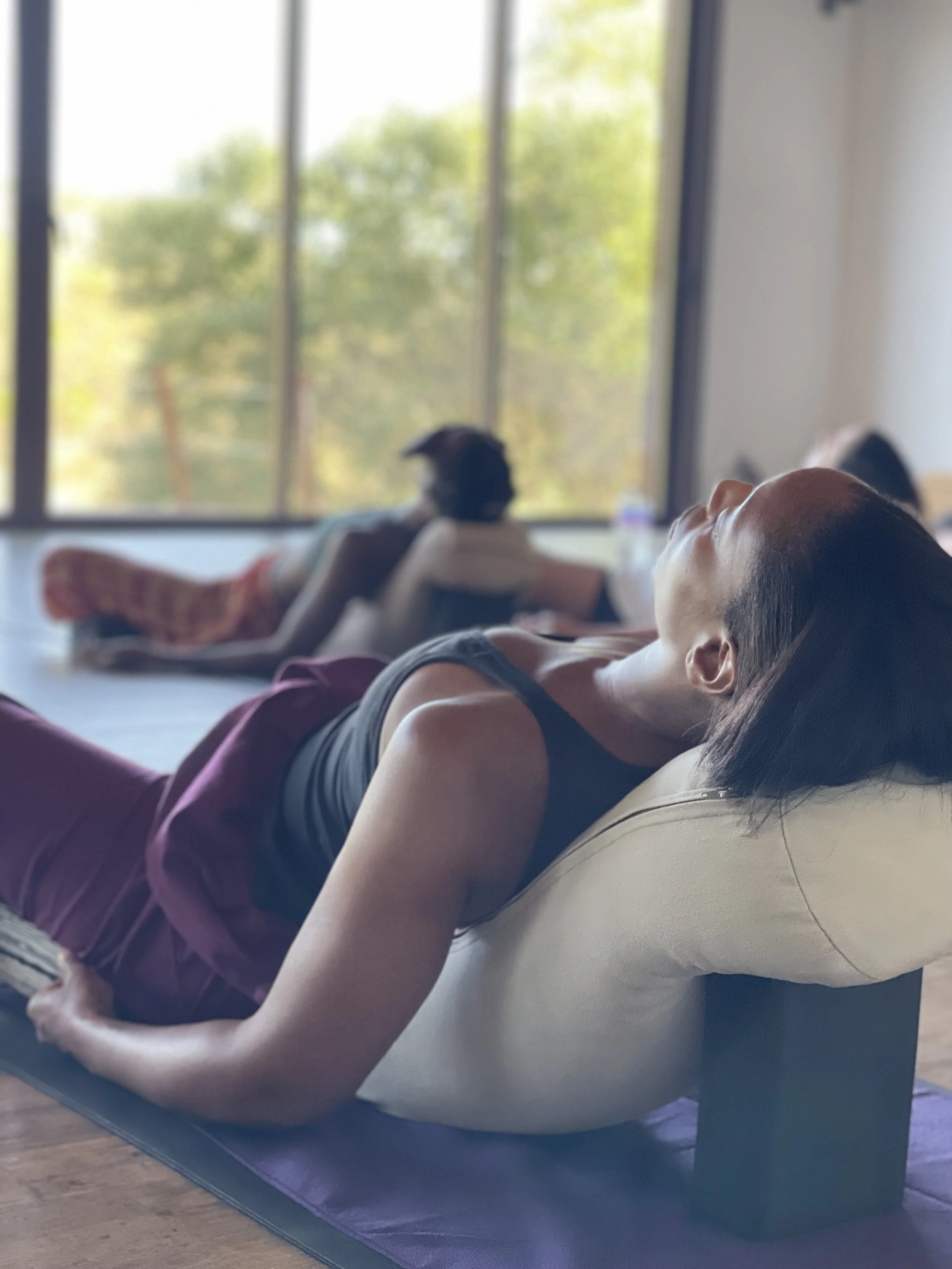 Black women in a yoga class lying on their back on yoga mats with a bolster, relaxing during meditation, with large windows showing green trees outside, on a yoga retreat in Portugal.