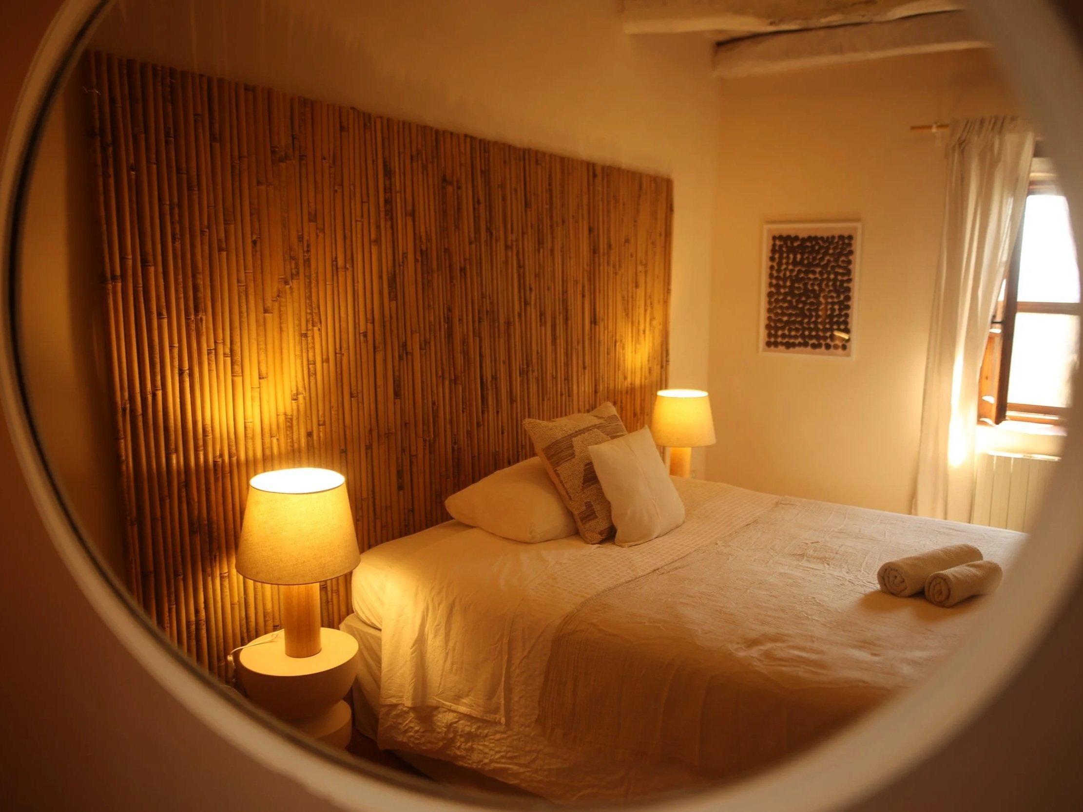 A cozy hotel room with a large bed, beige pillows, wood headboard, hanging woven lamp, curtains covering a balcony door, potted plant, and a white dresser with a kettle and cups.