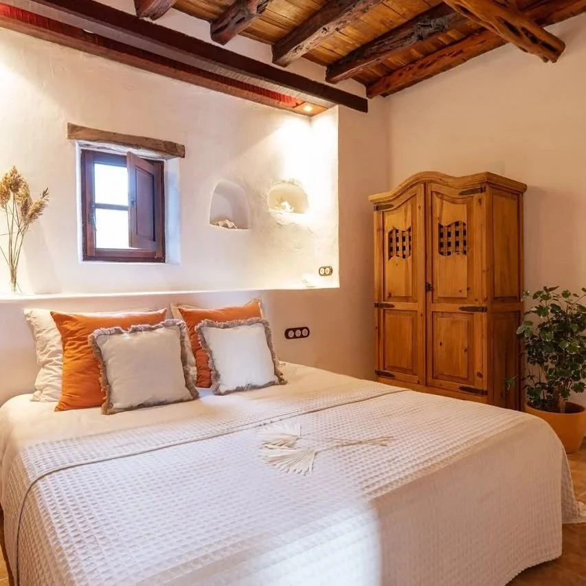 A cozy bedroom with a large bed, beige headboard, multiple pillows, bedside lamp, wooden side table, and sheer curtains allowing natural light.