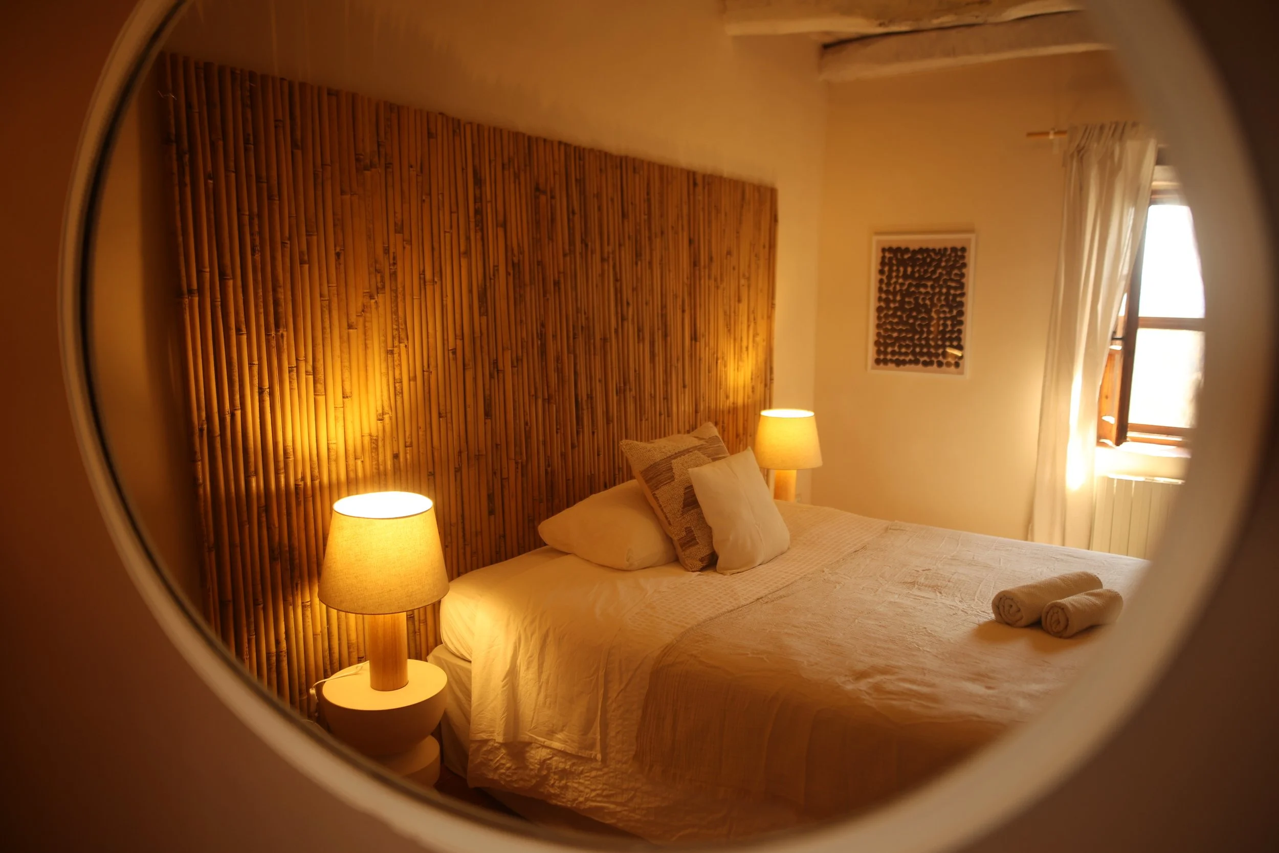 A cozy hotel room with a large bed, beige pillows, wood headboard, hanging woven lamp, curtains covering a balcony door, potted plant, and a white dresser with a kettle and cups.